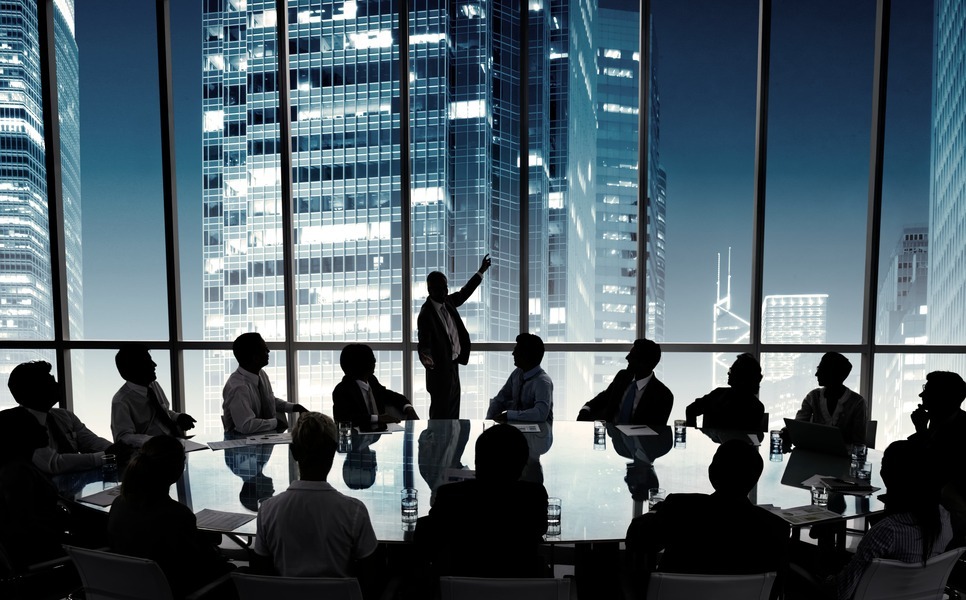 A formal business meeting in a modern boardroom with floor-to-ceiling windows overlooking a nighttime city skyline. A presenter stands and gestures while speaking to a group of seated professionals around a large conference table, suggesting a corporate discussion or strategic presentation