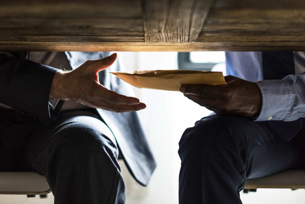 A person secretly passing an envelope under a table to another person, symbolizing corruption and bribery.
