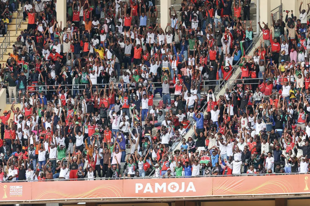 A vibrant crowd in a stadium raising their hands in unison, many dressed in red, with a visible "PAMOJA" banner symbolizing unity during the CHAN 2024 event in Kenya