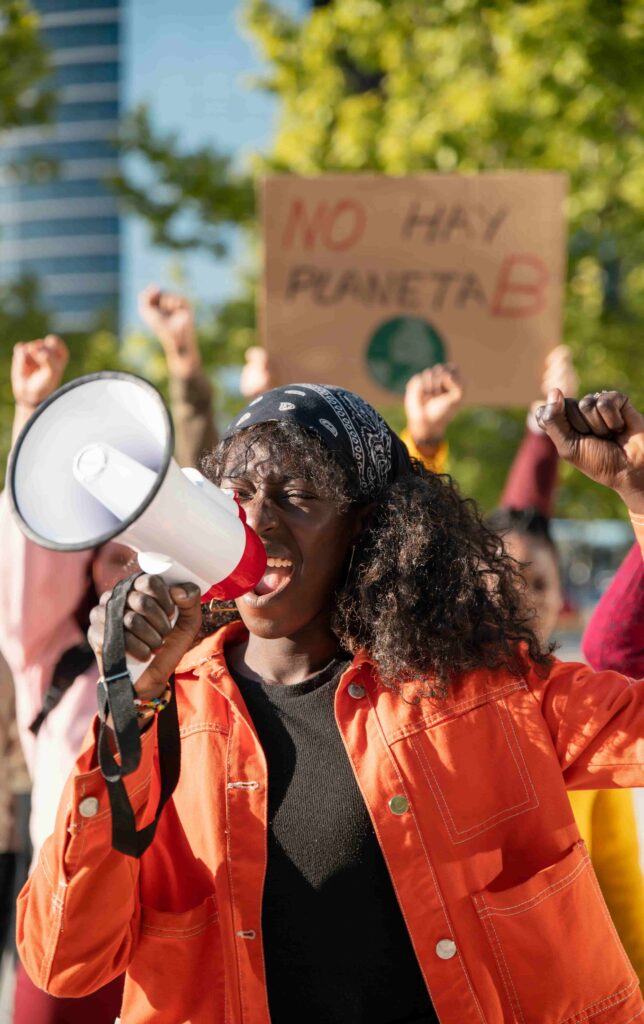 A protester speaking passionately through a megaphone while raising a fist during a climate change demonstration, with a visible placard in the background reading “No hay planeta B” (There is no planet B).