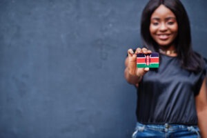 A person holding a small rectangular object with the flag of Kenya, featuring black, red, green stripes and a white border with a Maasai shield and spears in the center.