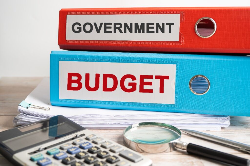 Two labeled binders stacked on a desk, one red with the word 'Government' and one blue with the word 'Budget', alongside a calculator, magnifying glass, and pen on top of scattered papers.