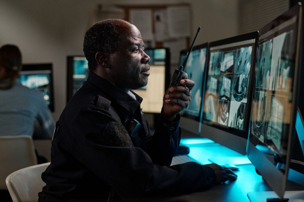 A security guard sitting in front of multiple surveillance monitors, speaking into a walkie-talkie while observing live camera footage in a control room.