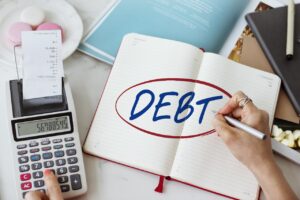 Person writing the word debt in a notebook with a calculator and receipt nearby, symbolizing financial planning and debt management.