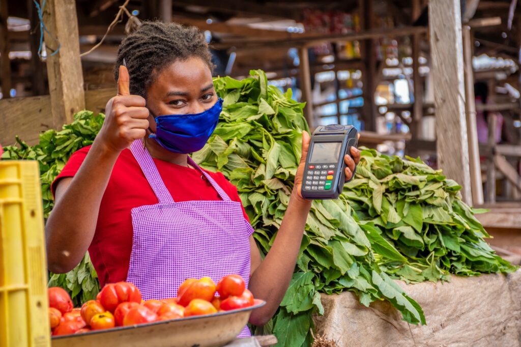 A masked vendor in a red shirt and purple checkered apron stands at a market stall, holding a card reader and giving a thumbs-up beside trays of tomatoes and leafy vegetables.