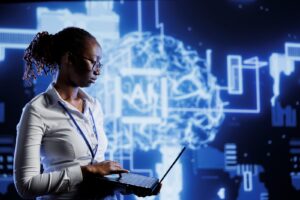 A person in glasses and a white shirt works on a laptop in front of a glowing digital brain illustration with circuitry and the letters AI, symbolizing artificial intelligence and technology.