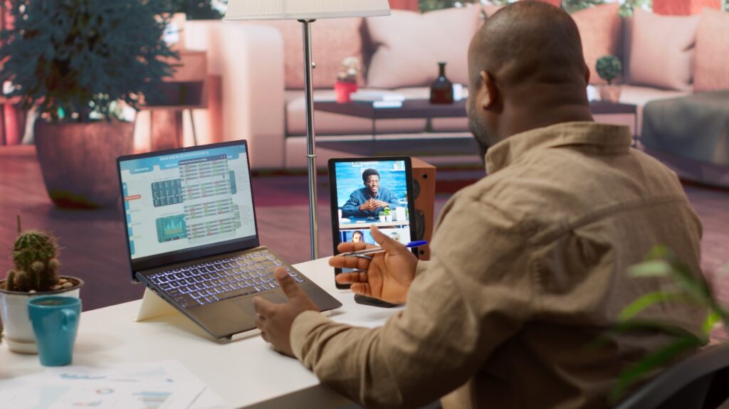 Person sitting at a desk during a video call, with a laptop showing charts under the heading “MAIN HUB OF STATISTICS & DATA,” a tablet displaying another participant, a blue mug, cactus plant, and printed charts, in a modern living room setting.