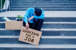 Person in a blue shirt and black tie sitting on outdoor concrete steps with a cardboard box of office belongings, holding a sign that reads “LOOKING FOR A JOB,” symbolizing unemployment and job searching.